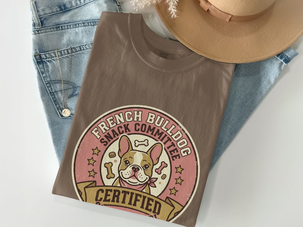 Brown t-shirt with French bulldog and "Snack Committee" text, laid on jeans and hat.