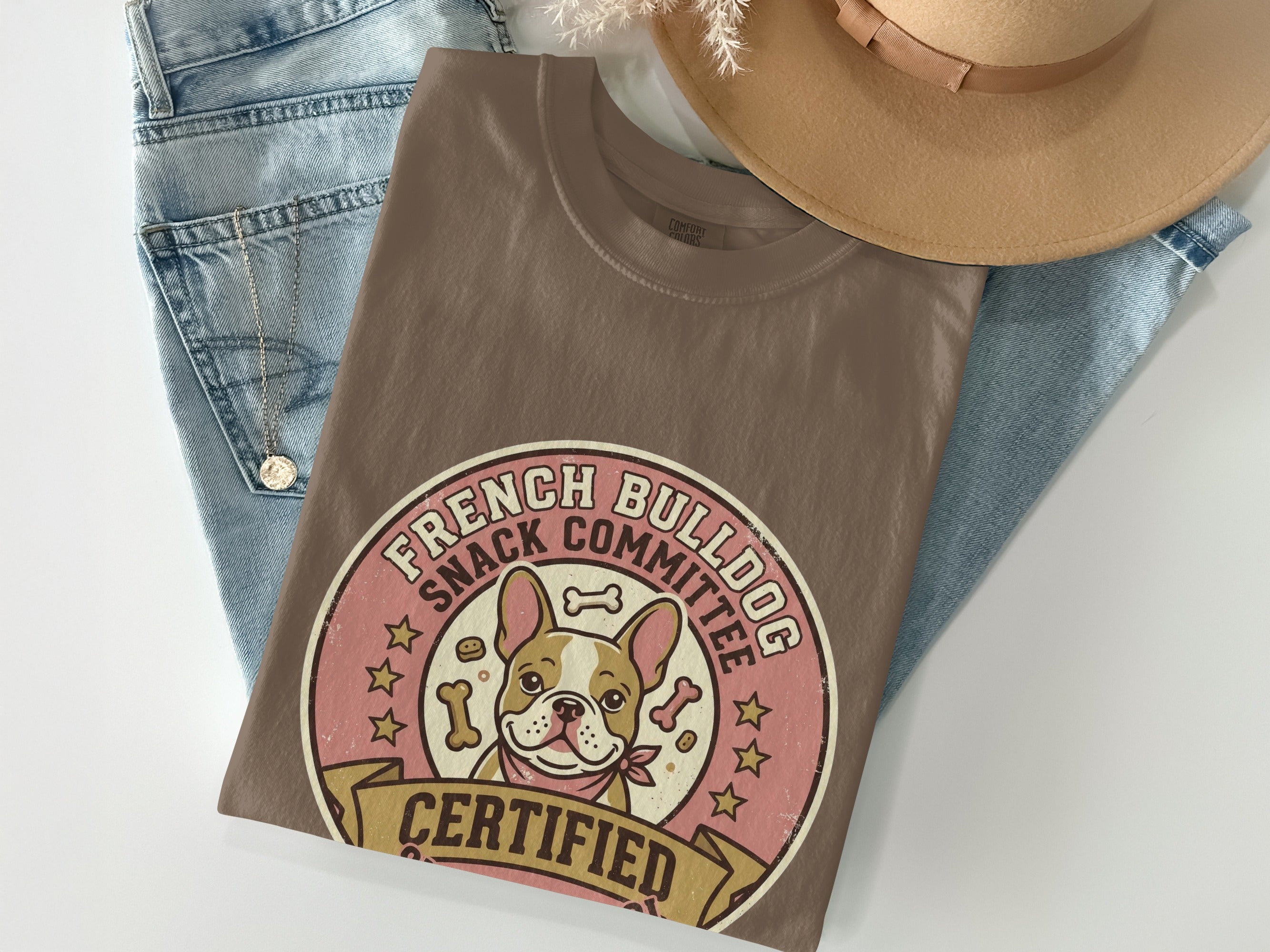 Brown t-shirt with French bulldog and "Snack Committee" text, laid on jeans and hat.